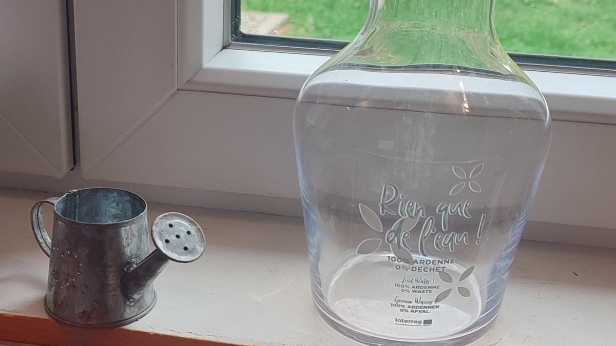 La carafe « Rien que de l’eau ! 100 % Ardenne, 0 % déchet » sur le rebord de la fenêtre de la cuisine, devant le jardin.