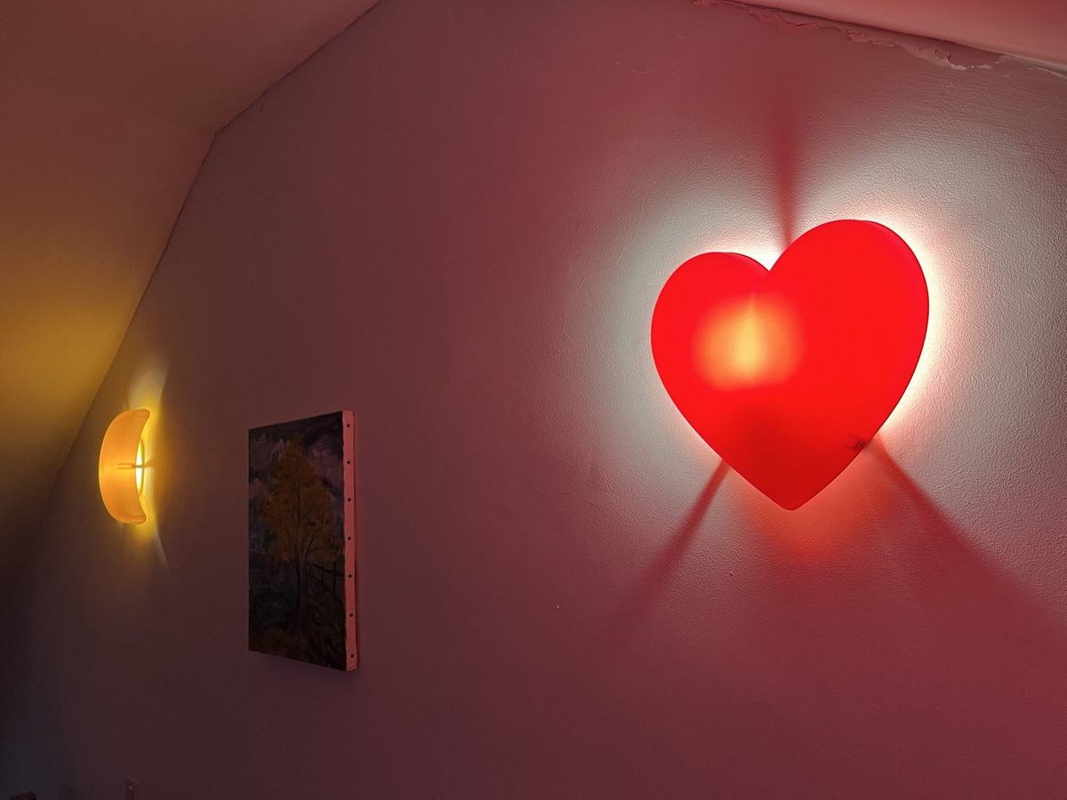 Le mur de la chambre des enfants : une applique en forme de cœur rouge lumineux, une applique en forme de lune orange et une petite toile peinte d’un arbre en automne.