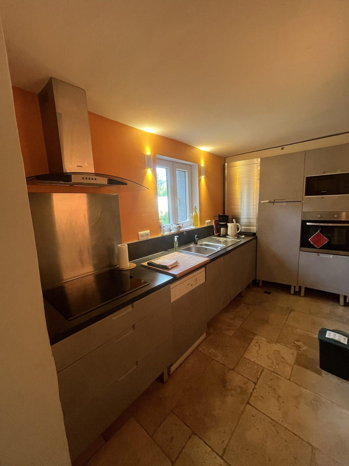 La cuisine équipée : plaque à induction, hotte inox, lave-vaisselle, four et micro-ondes en colonne, mur orange et sol en pierre.