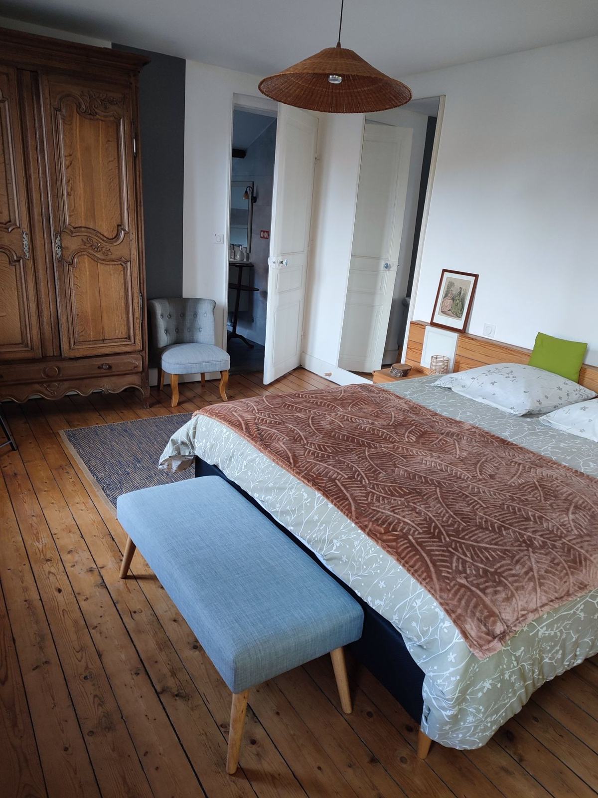 La chambre du premier étage : armoire ancienne en chêne, fauteuil crapaud gris, suspension en rotin et lit deux personnes sur le parquet.