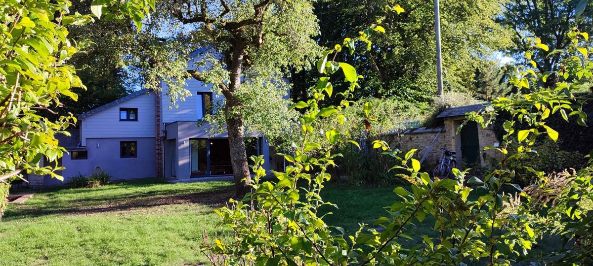 La maison vue du fond du jardin en fin de journée : pelouse, grands arbres, baies vitrées ouvertes et l’abri à vélos contre le mur de pierre.