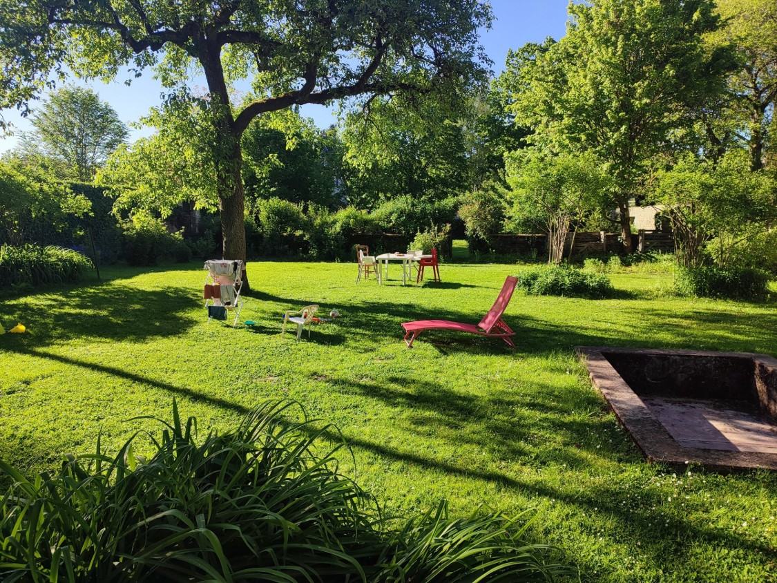 Le jardin au printemps : pelouse sous un grand arbre, bain de soleil rouge, table et chaises de jardin, étendoir à linge.