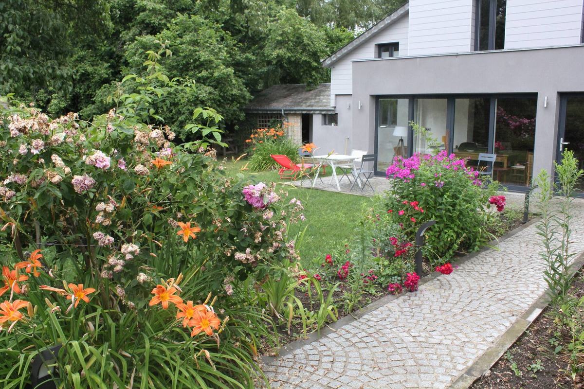 Le jardin fleuri du gîte en été : hémérocalles orange et rosiers au premier plan, allée pavée menant à la terrasse et aux baies vitrées de la maison.