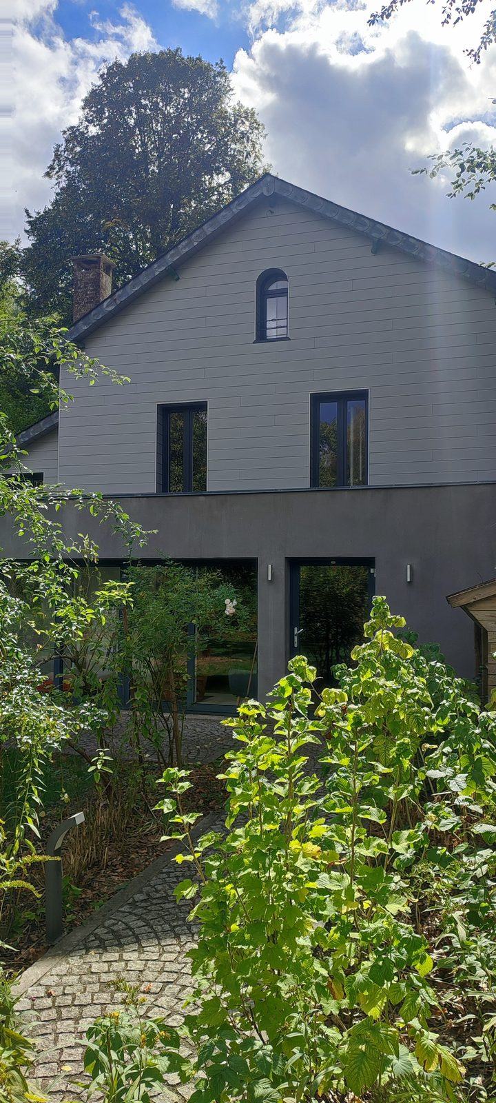 La façade de la maison vue du jardin : bardage clair, extension vitrée de plain-pied et allée pavée.