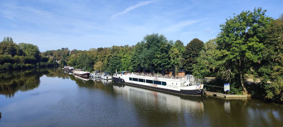 La Meuse vue d’en haut, une péniche-restaurant et des bateaux amarrés le long des rives boisées.