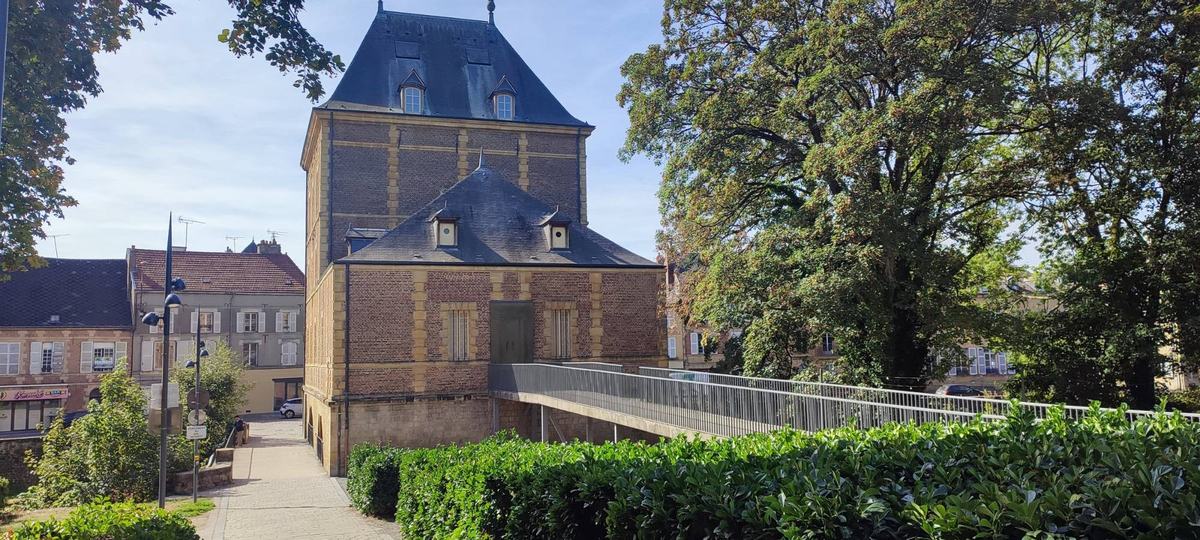 Le Vieux Moulin, qui abrite le musée Rimbaud, en brique et pierre, avec la passerelle qui y mène et une haie au premier plan.