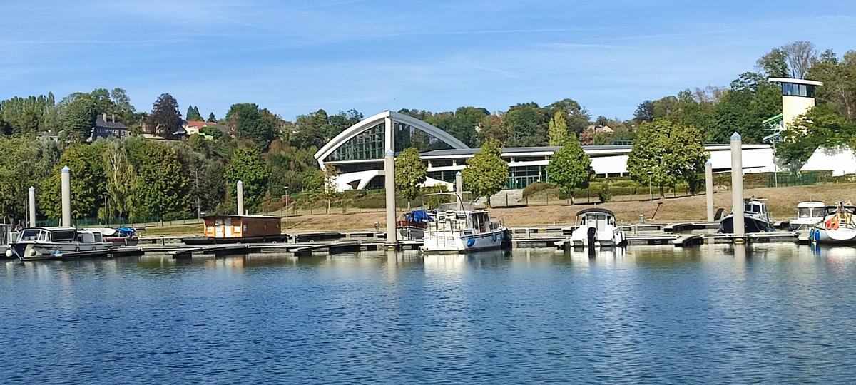 Le port de plaisance sur la Meuse : pontons et bateaux, le centre aquatique et son toit courbe sur la rive d’en face.