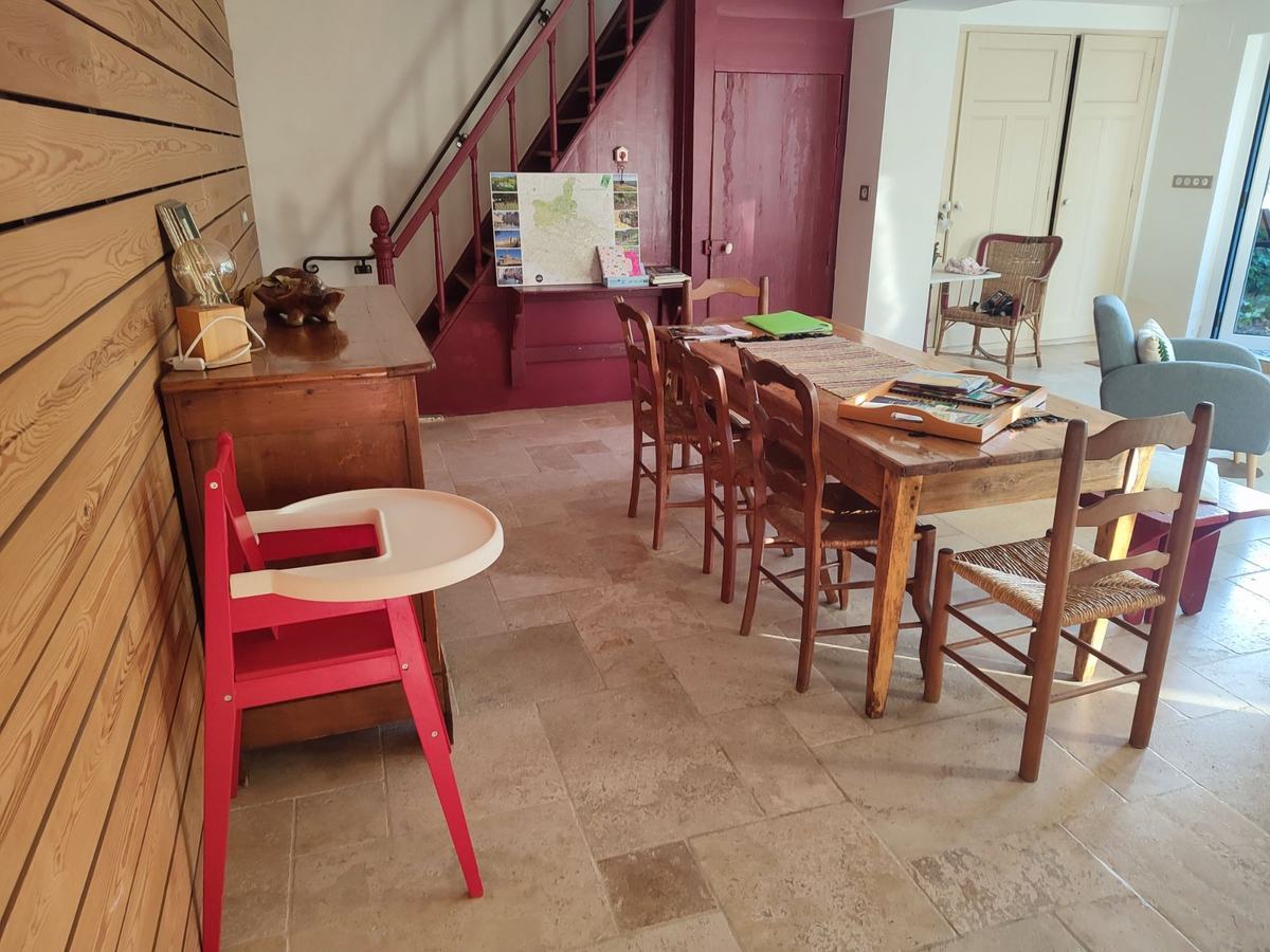 La salle à manger : grande table en bois et chaises paillées, chaise haute rouge, escalier framboise et carte de la région au mur.