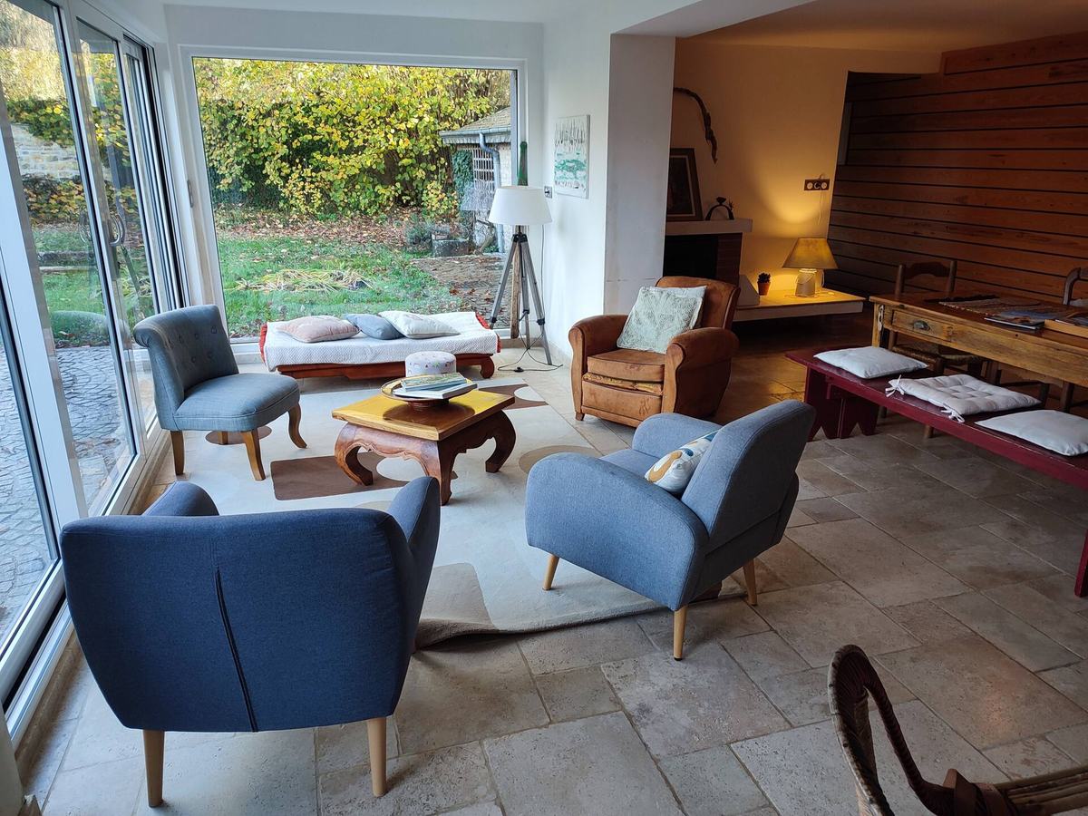 Le salon : fauteuils bleus autour d’une table basse en bois, sol en travertin, baies vitrées ouvrant sur le jardin.