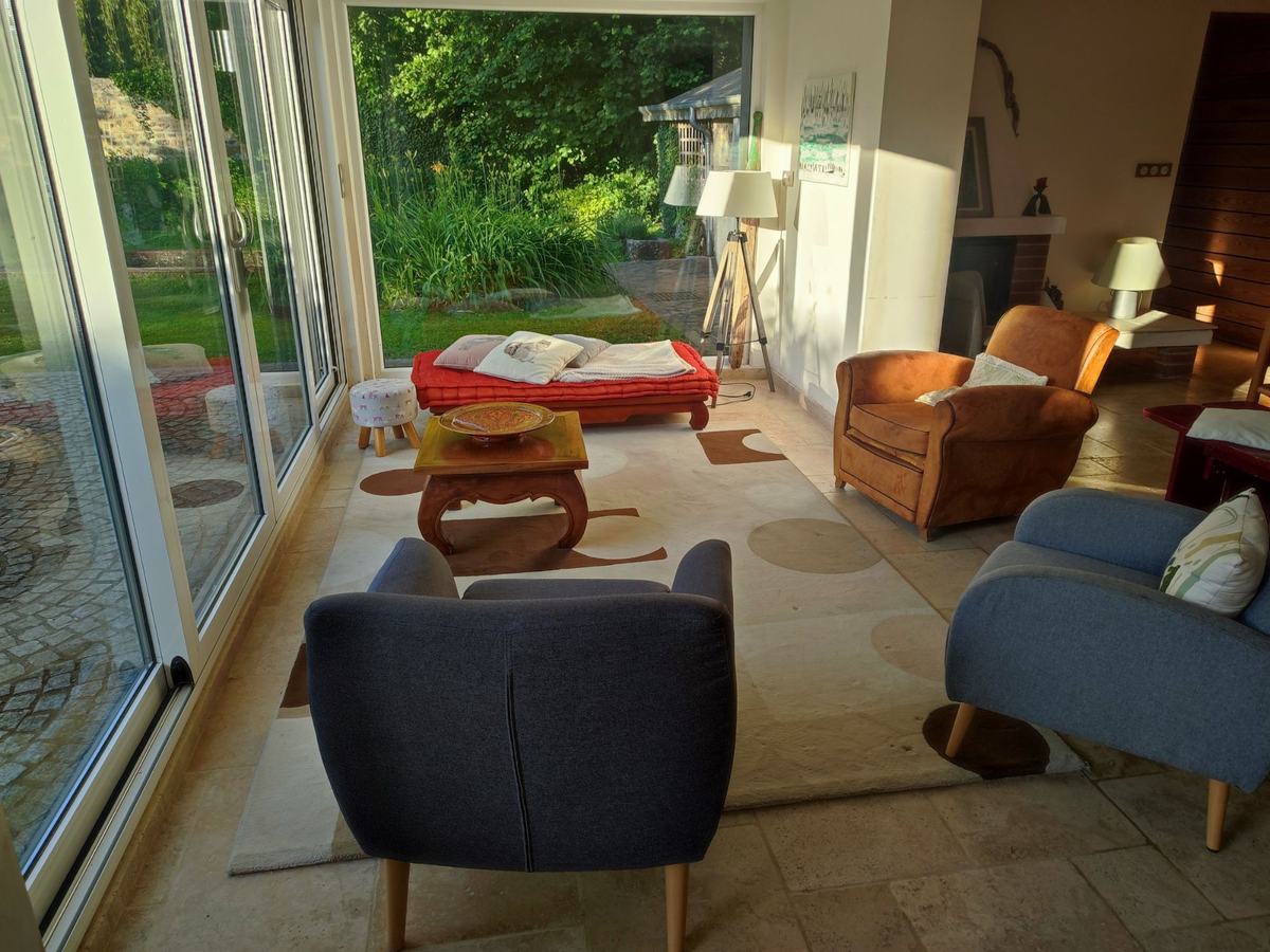 Le salon en fin de journée : fauteuils club en cuir, fauteuils gris, table basse et banquette rouge devant les baies vitrées ouvertes sur le jardin.