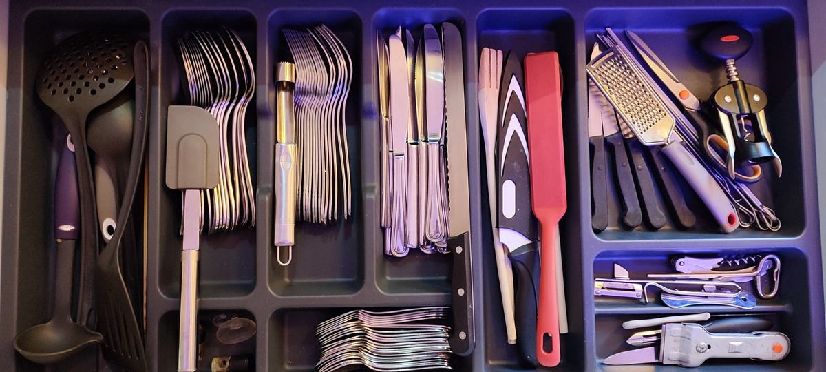 Le tiroir de la cuisine ouvert : couverts de table, couteaux de cuisine, couteau à pain, louche, écumoire, spatules, râpe, ciseaux, économe, ouvre-boîte et tire-bouchon, chacun dans son compartiment.