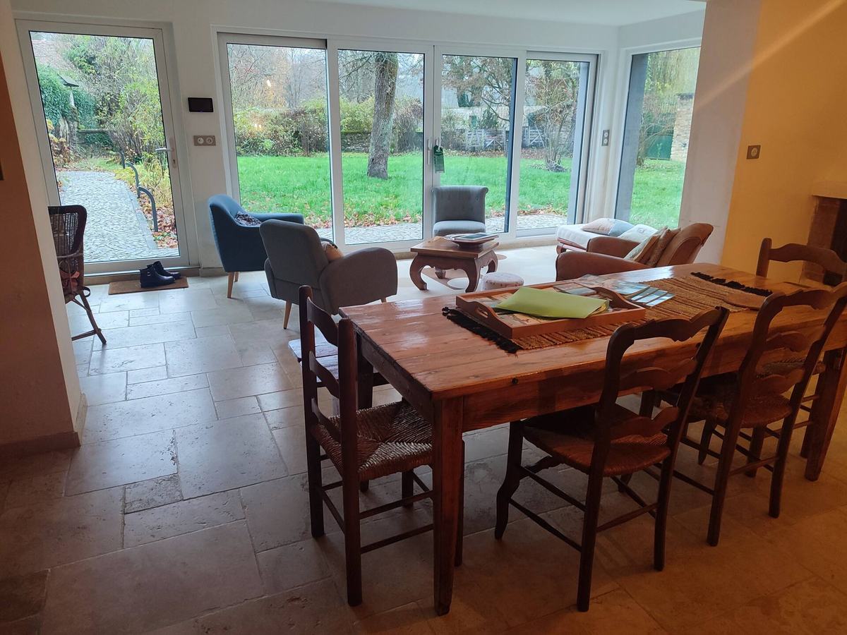 La véranda et la salle à manger : table en bois dressée, chaises anciennes, baies vitrées d’angle sur le jardin.