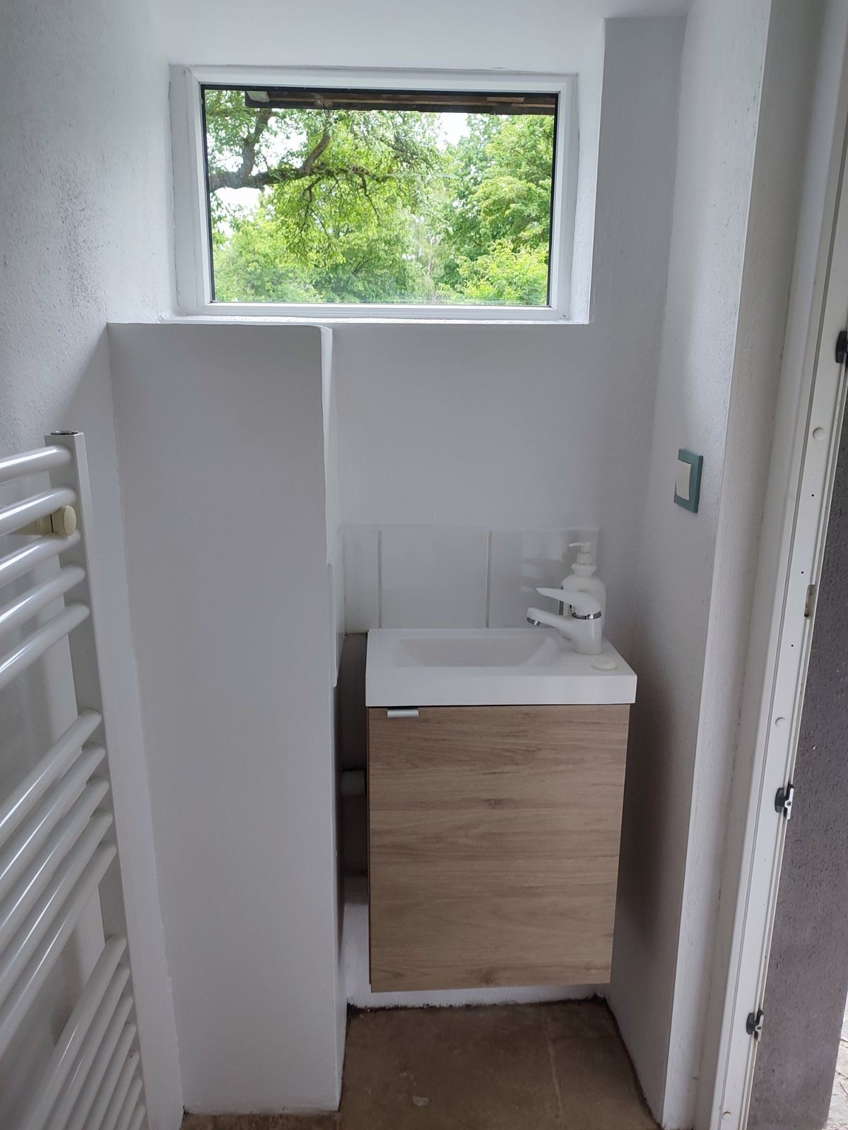 Le second WC, accessible de l’extérieur : lave-mains blanc sur meuble en bois, sèche-serviettes et fenêtre sur les arbres.